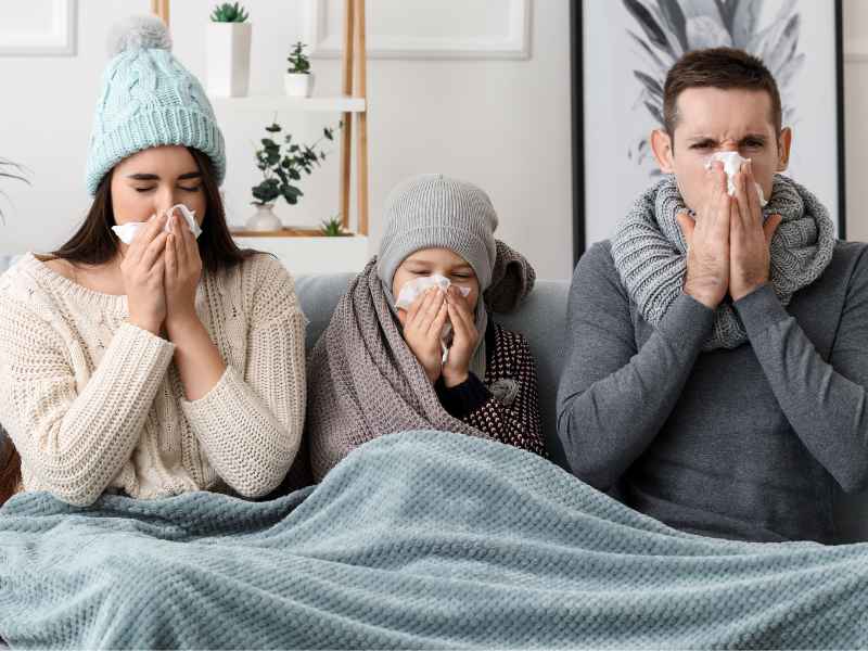 Family of three under a blanket on couch, all blowing their noses.